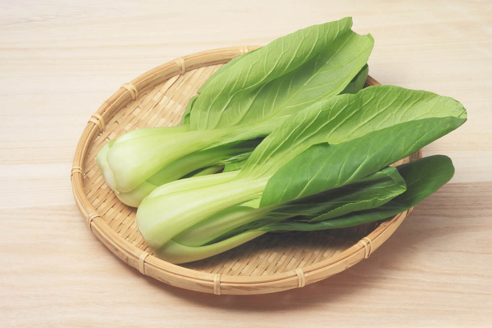 Two fresh bok choy stalks with green leaves on a round woven bamboo plate, placed on a light wooden surface.
