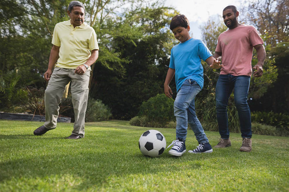 A boy in a blue shirt is playing soccer on a grassy lawn, while two adults, one in a yellow shirt and the other in a pink shirt, watch and interact with him.