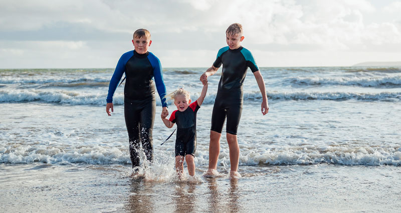 Two older boys in wetsuits are holding hands with a younger child in a swimsuit while standing in shallow water at the beach. The ocean waves are visible in the background.