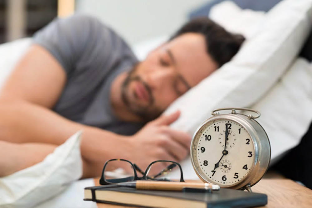 A man is sleeping on a bed with a relaxed expression. In the foreground, there is an alarm clock and a pair of glasses on a bedside table, indicating a cozy bedroom setting.