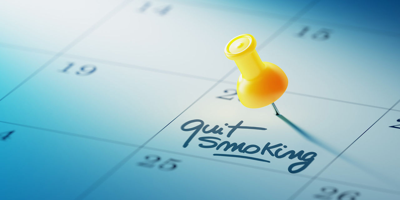 A yellow pushpin on a square of a blue calender that has "quit smoking" written and underlined within. 