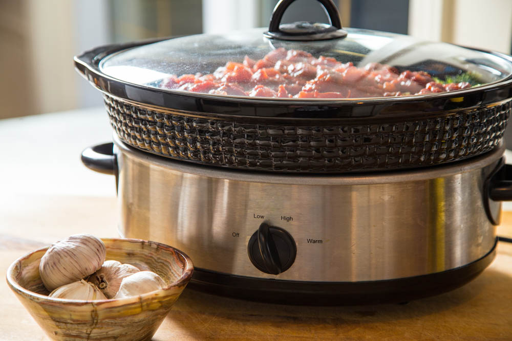Slow cooker with a glass lid filled with simmering food, placed on a kitchen counter next to a bowl of garlic.