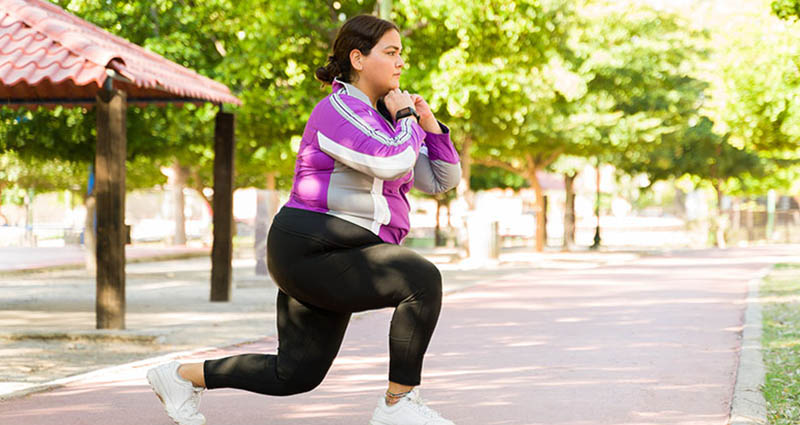A woman is exercising outdoors, performing a lunge on a pathway surrounded by trees. She is wearing a purple and white top with black leggings, indicating an active lifestyle.