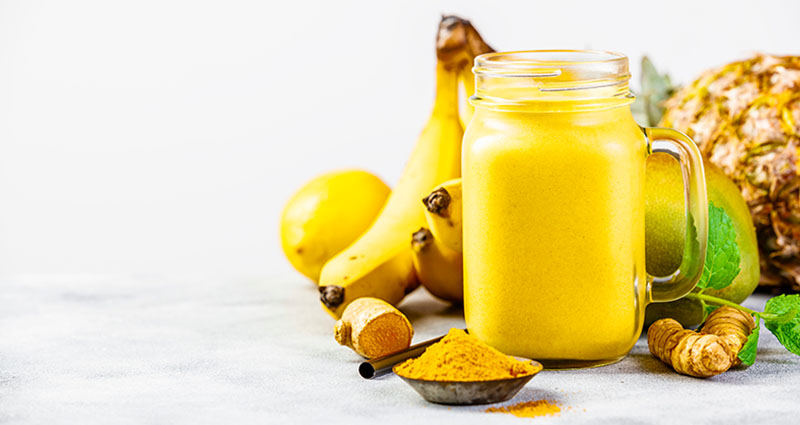 A jar of yellow smoothie with bananas, pineapple, ginger, turmeric powder, and mint leaves on a light surface.