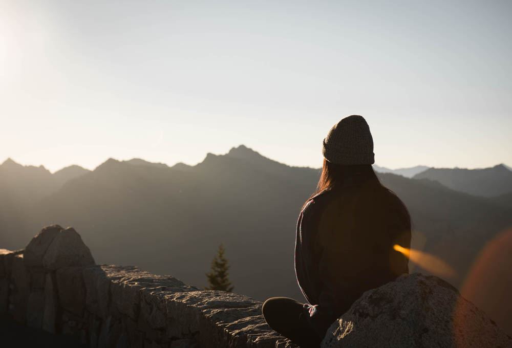 Person in a beanie sitting on a stone wall, facing mountains under a clear sky at sunrise or sunset.