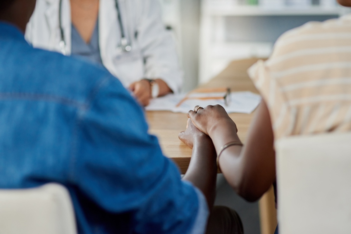 Two people sitting at a table holding hands while talking to a doctor in a white coat, who is partially visible in the background.