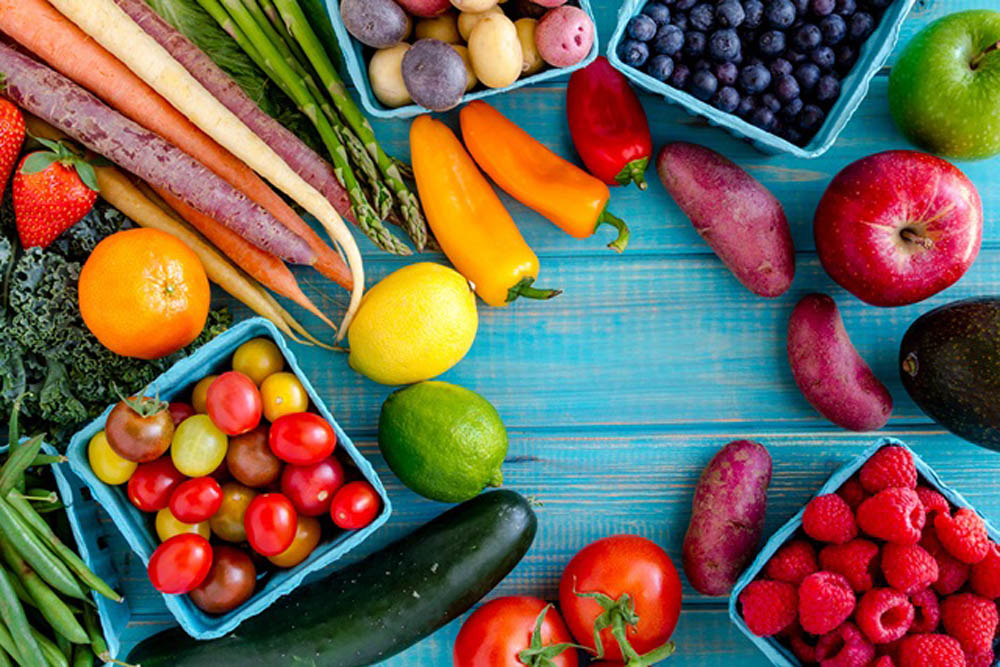 An overhead view of a variety of fresh fruits and vegetables arranged on a blue wooden surface. Items include tomatoes, apples, carrots, peppers, and berries, showcasing a colorful assortment of produce.