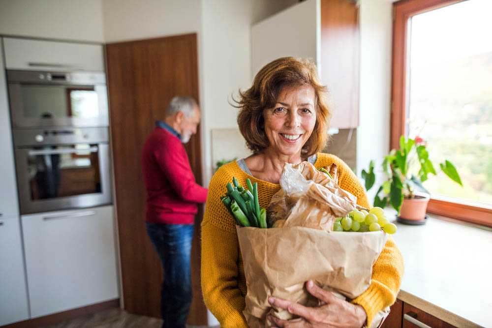 A woman in a kitchen holding a paper bag with vegetables and grapes with a man in the background standing near a countertop.