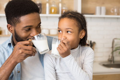 A father and daughter are smiling at each other while enjoying glasses of milk in a cozy kitchen.