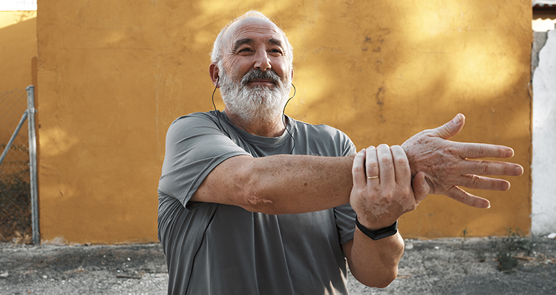 An older man with a beard is stretching his arms outdoors against a vibrant orange wall. He appears focused and engaged in a warm-up exercise.