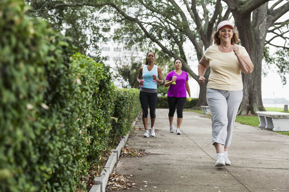 Three women walking on a tree-lined path, two holding water bottles in exercise attire; one leads, wearing a cap and glasses.