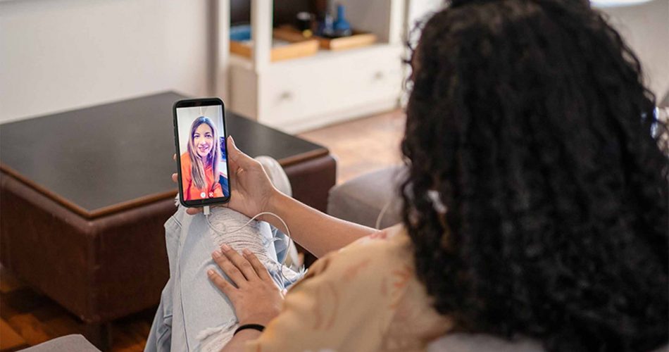 A person is sitting on a couch, holding a smartphone that displays a video call. The individual appears engaged and smiling, suggesting a friendly conversation. The room has a cozy atmosphere.