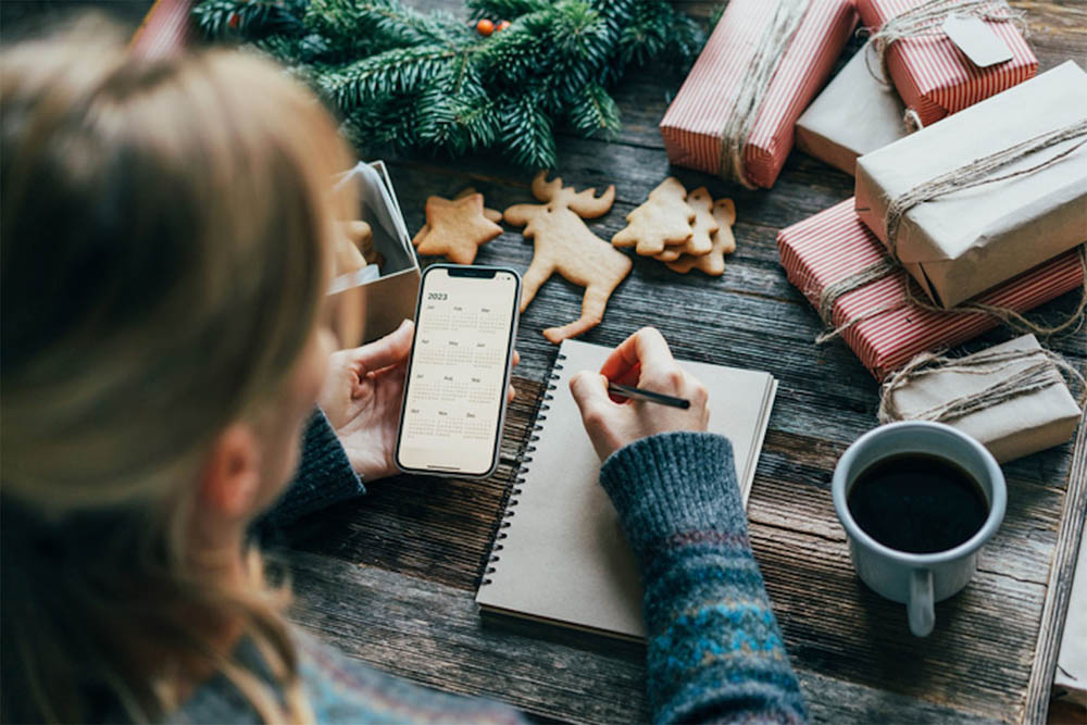 A person is writing in a notebook while surrounded by holiday decorations, gift-wrapped presents, and a cup of coffee. The scene conveys a cozy and festive atmosphere.