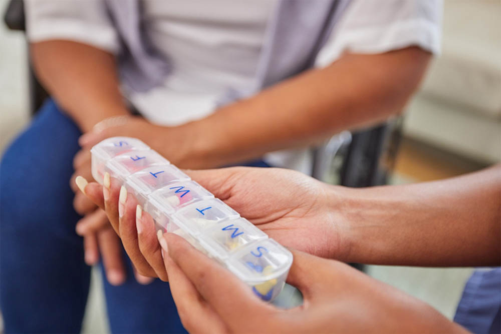  close-up of hands holding a weekly pill organizer, with clearly labeled compartments for each day. The background shows another person sitting nearby, suggesting a setting focused on health management.