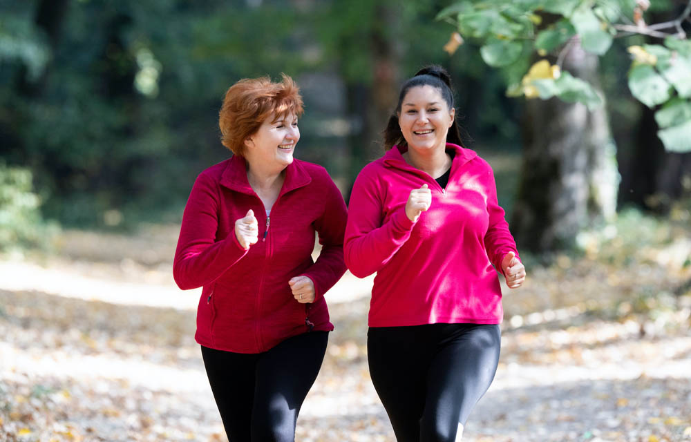 Two women in red jackets jogging on a forest path, surrounded by trees and fallen leaves.