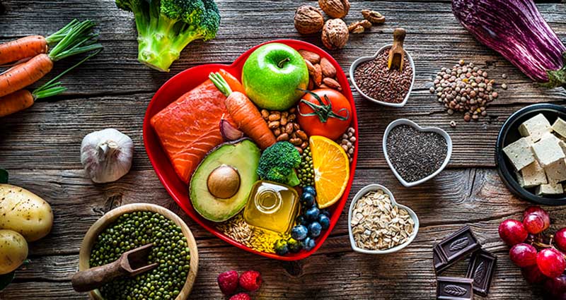 An assortment of fresh foods arranged in a heart-shaped container, including salmon, fruits, vegetables, nuts, and seeds, displayed on a rustic wooden surface.