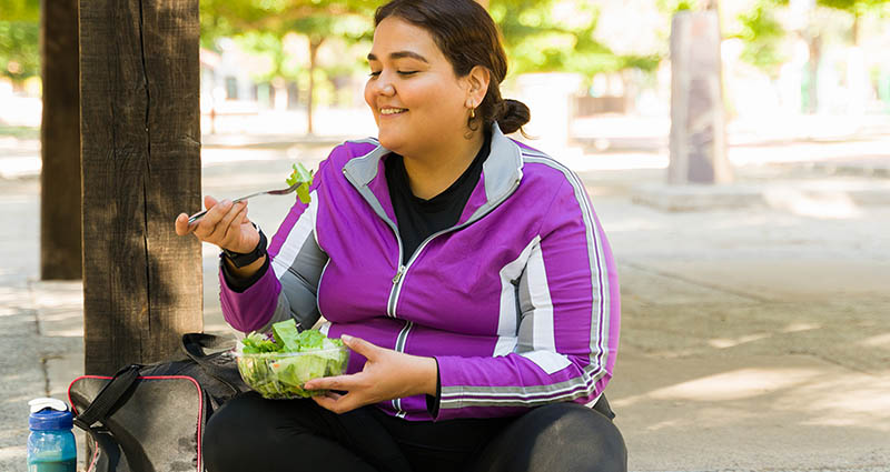A woman in a purple jacket is enjoying a salad while sitting outdoors, smiling contentedly. The environment is bright and green, suggesting a pleasant day.