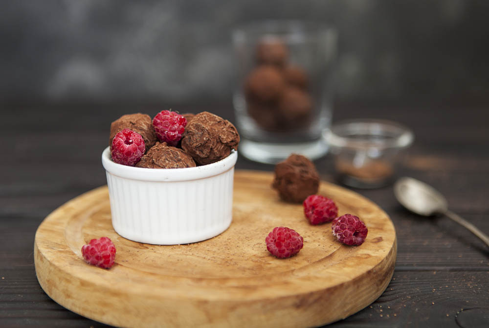 A white ramekin filled with chocolate truffles and raspberries is on a wooden board, with additional truffles, raspberries, and a spoon nearby.