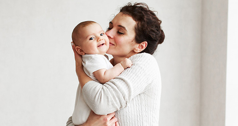 A woman is lovingly holding a baby and giving them a kiss on the cheek, both smiling. The setting is warm and intimate, highlighting their affectionate bond.