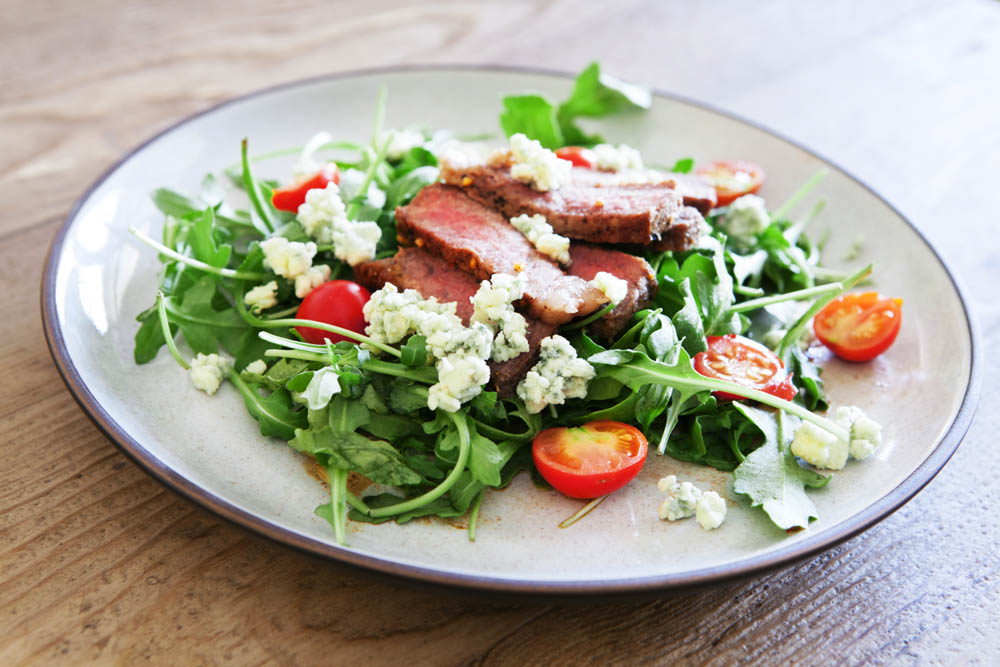 Salad with sliced steak, arugula, cherry tomatoes, and crumbled blue cheese on a white plate, placed on a wooden table.