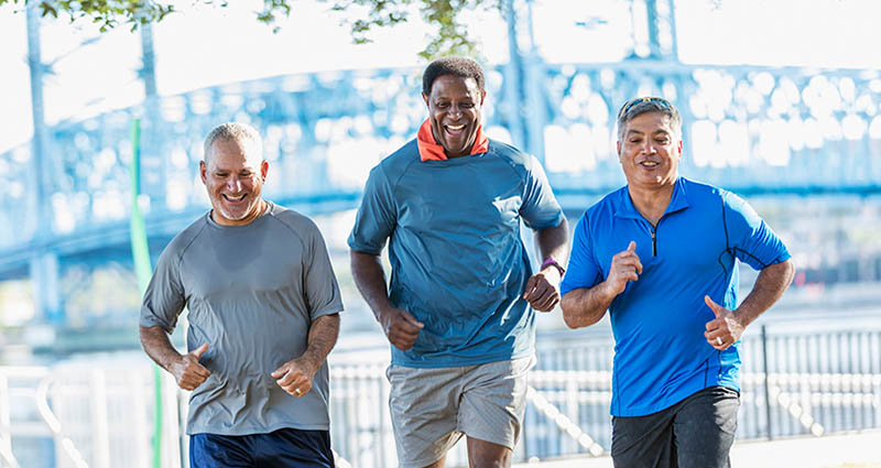  Three men are jogging together outdoors, smiling and enjoying their exercise. They are dressed in athletic clothing, with a bridge visible in the background.