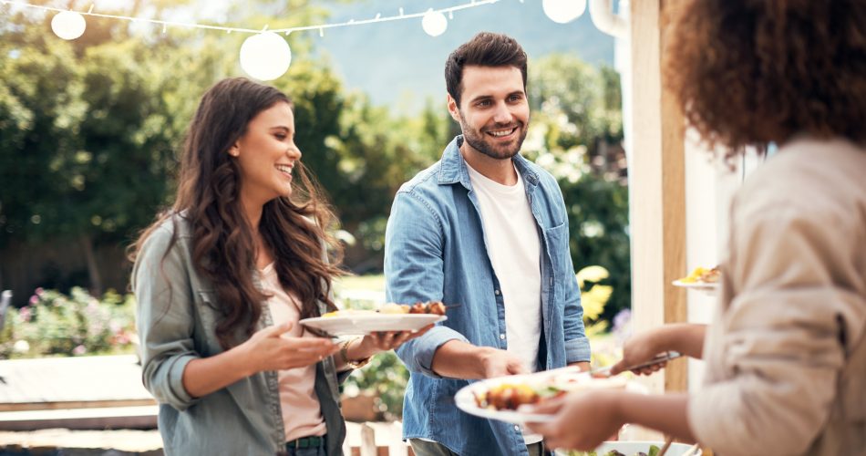 A man and woman are smiling and holding plates of food while interacting with another person outdoors, likely at a social gathering. The setting is bright and cheerful with string lights in the background.