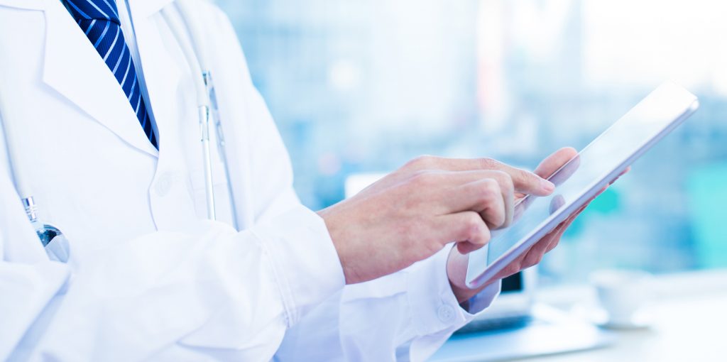 A close-up of a person in a white lab coat using a tablet, likely in a medical or professional setting. The background is blurred, focusing on the interaction with the device.