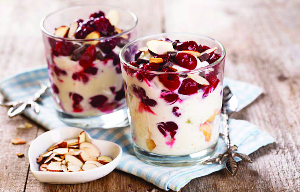 Two glasses of yogurt topped with red berries and almond slices, placed on a blue checkered cloth, with a small bowl of almonds beside them.