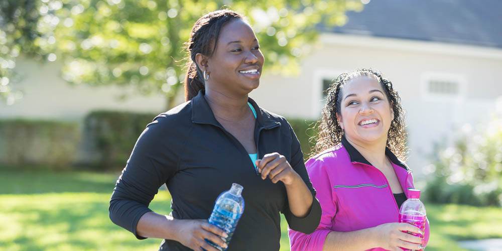  Two women are walking outdoors with water bottles, smiling and enjoying each other's company in a sunny environment.