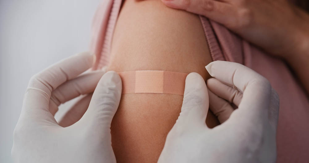 Gloved hands applying a bandage to a persons upper arm, suggesting recent medical care or vaccination.