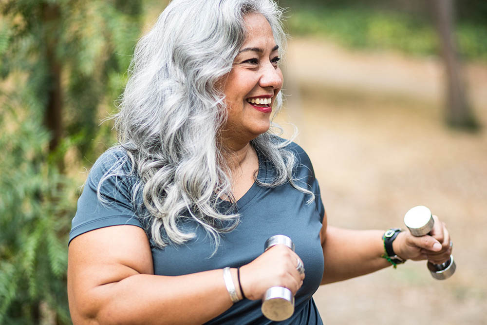  A woman with long, gray hair is smiling while holding dumbbells outdoors, indicating an active and healthy lifestyle.