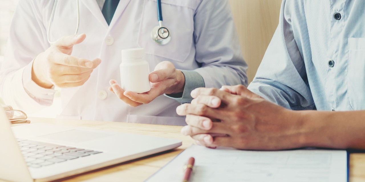 A doctor holding a white pill bottle, discussing it with a seated patient. A laptop and clipboard are on the table.