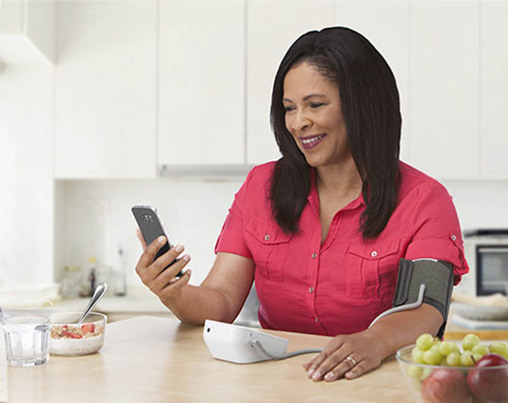  A woman in a red shirt checks her smartphone while wearing a blood pressure monitor at a kitchen table, with fruits in the background.