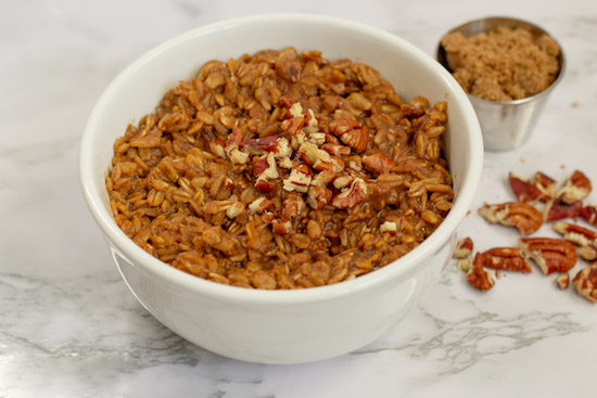 A white bowl filled with oatmeal topped with chopped pecans, with a small cup of brown sugar and scattered pecans on the side on a marble surface.
