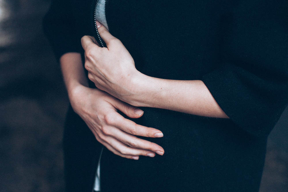 Person in a dark sweatshirt with hands held over their stomach, standing against a blurred background.