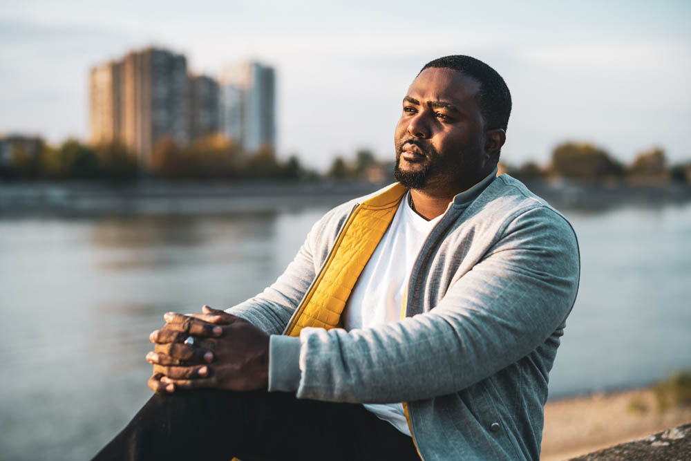 Man sitting outdoors near water, wearing a gray jacket and white shirt, with city buildings in the background.