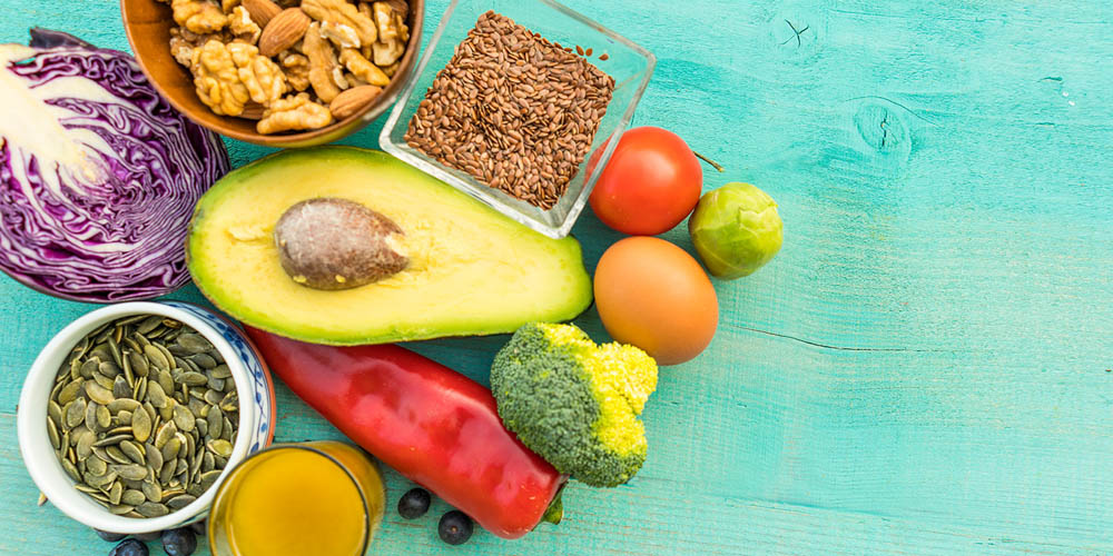 Assorted healthy foods including avocado, nuts, seeds, vegetables, and eggs on a turquoise wooden background.