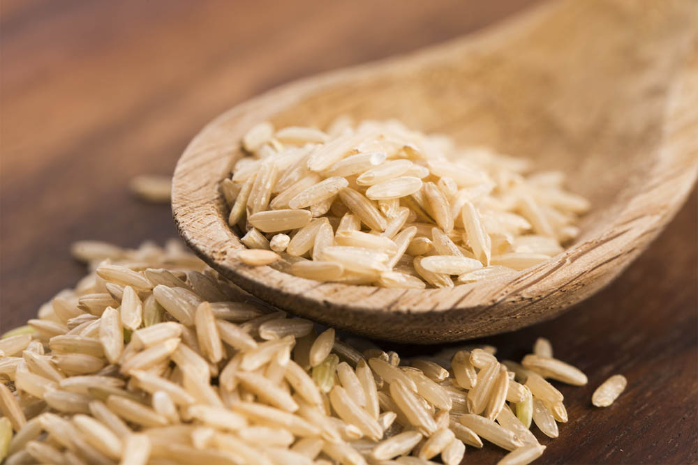 A wooden spoon filled with uncooked brown rice, resting on a wooden surface.