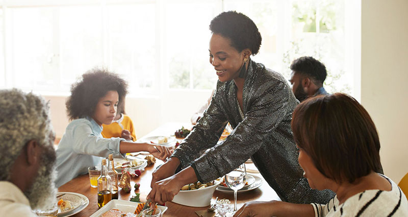 A family gathered around a table enjoying a meal together, with a woman serving food while smiling.