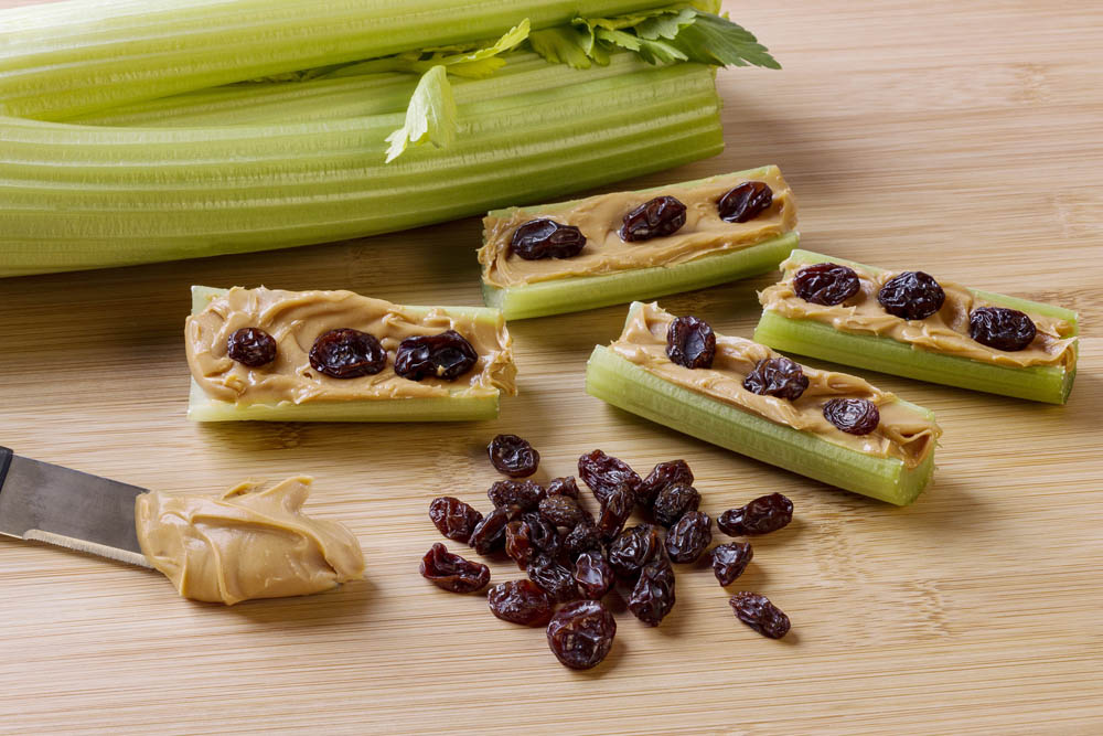 Celery sticks filled with peanut butter and topped with raisins, arranged on a wooden cutting board, with a knife and some extra peanut butter nearby.