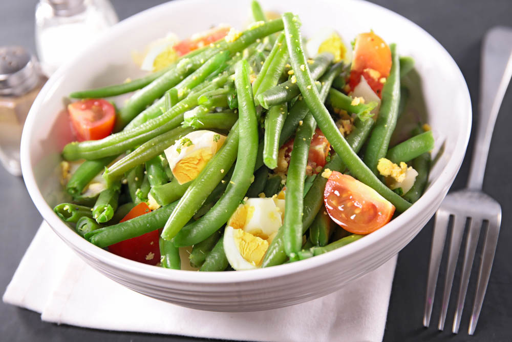 A white bowl filled with green beans, halved cherry tomatoes, and sliced boiled eggs on a white napkin with a fork nearby.