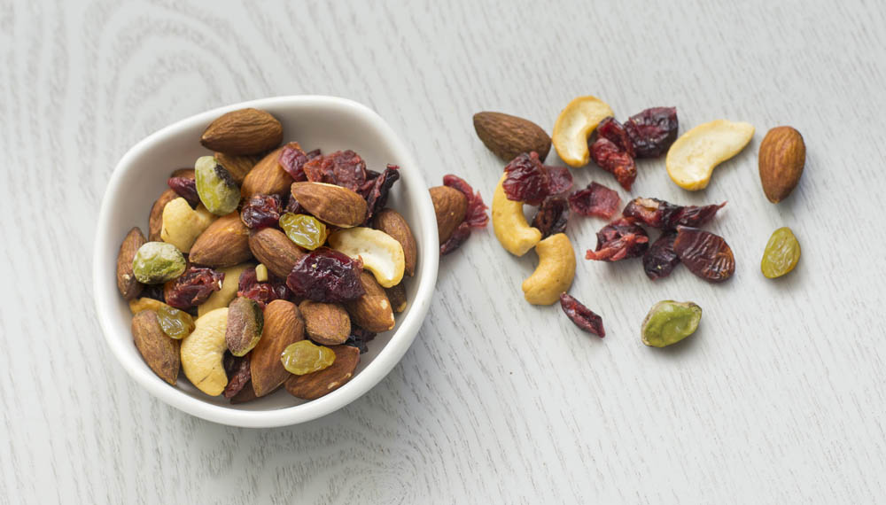 A bowl of mixed nuts and dried fruits, including almonds, cashews, raisins, and cranberries, with some scattered on a light wood surface.