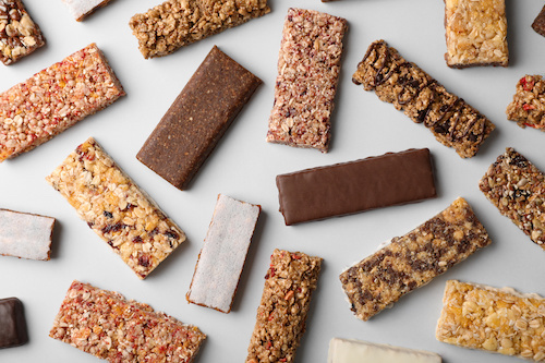 Various granola and energy bars with different textures and toppings scattered on a light gray surface.