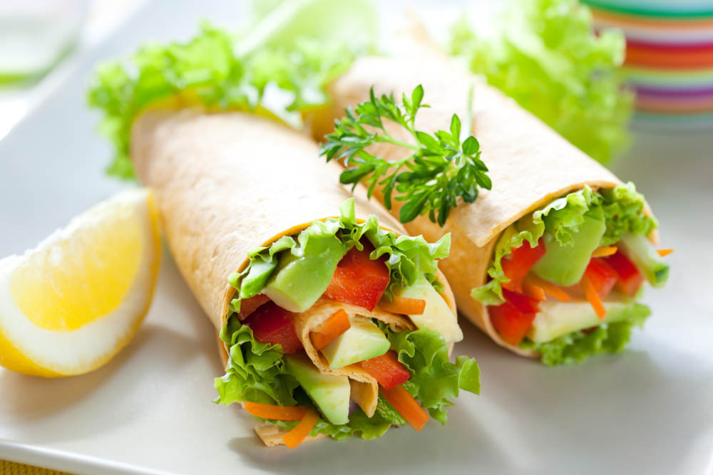 Two fresh vegetable wraps filled with lettuce, carrots, and cucumber, served on a white plate with a lemon wedge and garnished with parsley, with colorful bowls in the background.