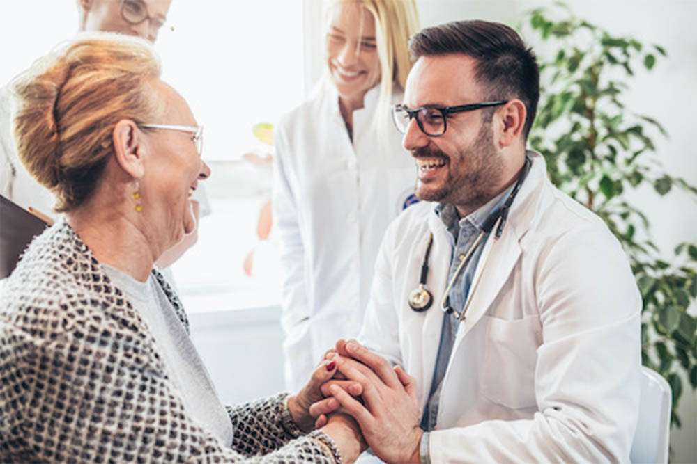 An elderly woman and a doctor smiling and holding hands during a consultation.