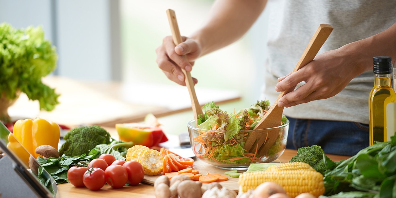 Person preparing a fresh salad in a bowl surrounded by colorful vegetables on a kitchen counter.