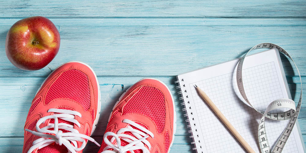 Red sneakers, a red apple, a notebook with a pencil, and a measuring tape on a blue wooden surface.