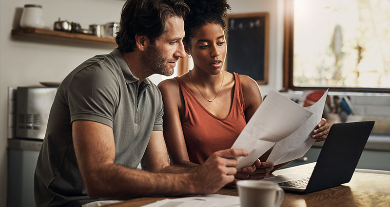 Couple at kitchen table reviewing documents with a laptop open in front of them