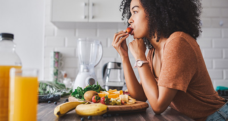 Young woman in a kitchen enjoying a fresh strawberry, surrounded by a variety of healthy fruits and juices.