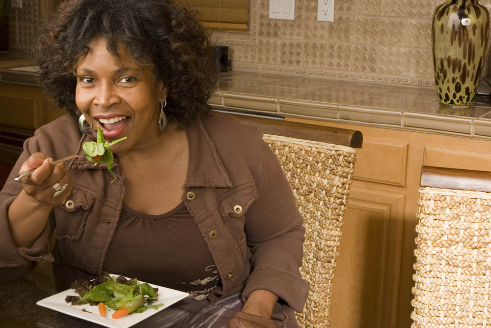 Woman smiling, seated at a kitchen counter, eating a salad. She holds a fork with greens near her mouth.